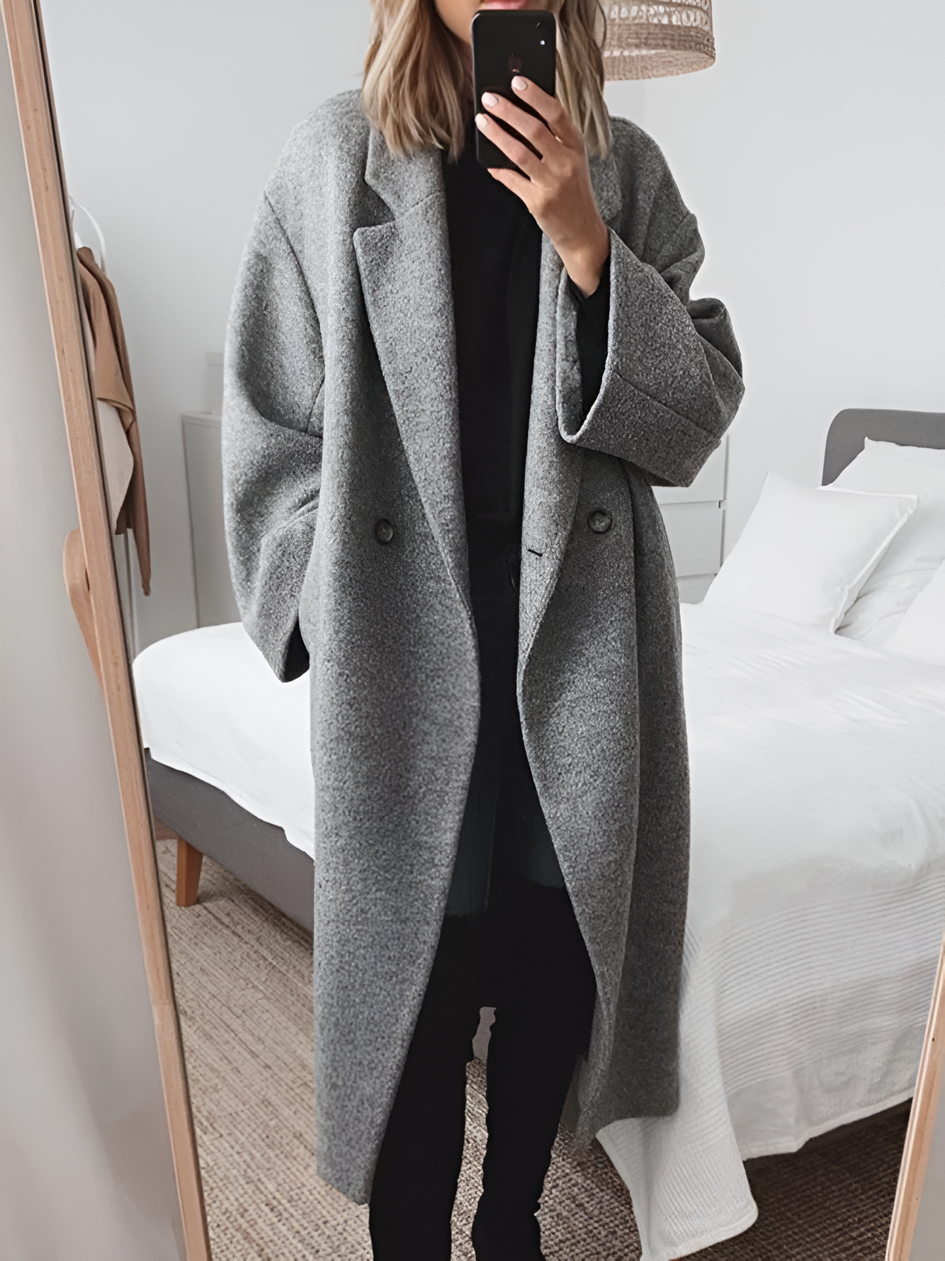 Femme en manteau gris oversize, prenant un selfie dans une chambre moderne. Mode automne-hiver, style minimaliste, tendance vestimentaire.