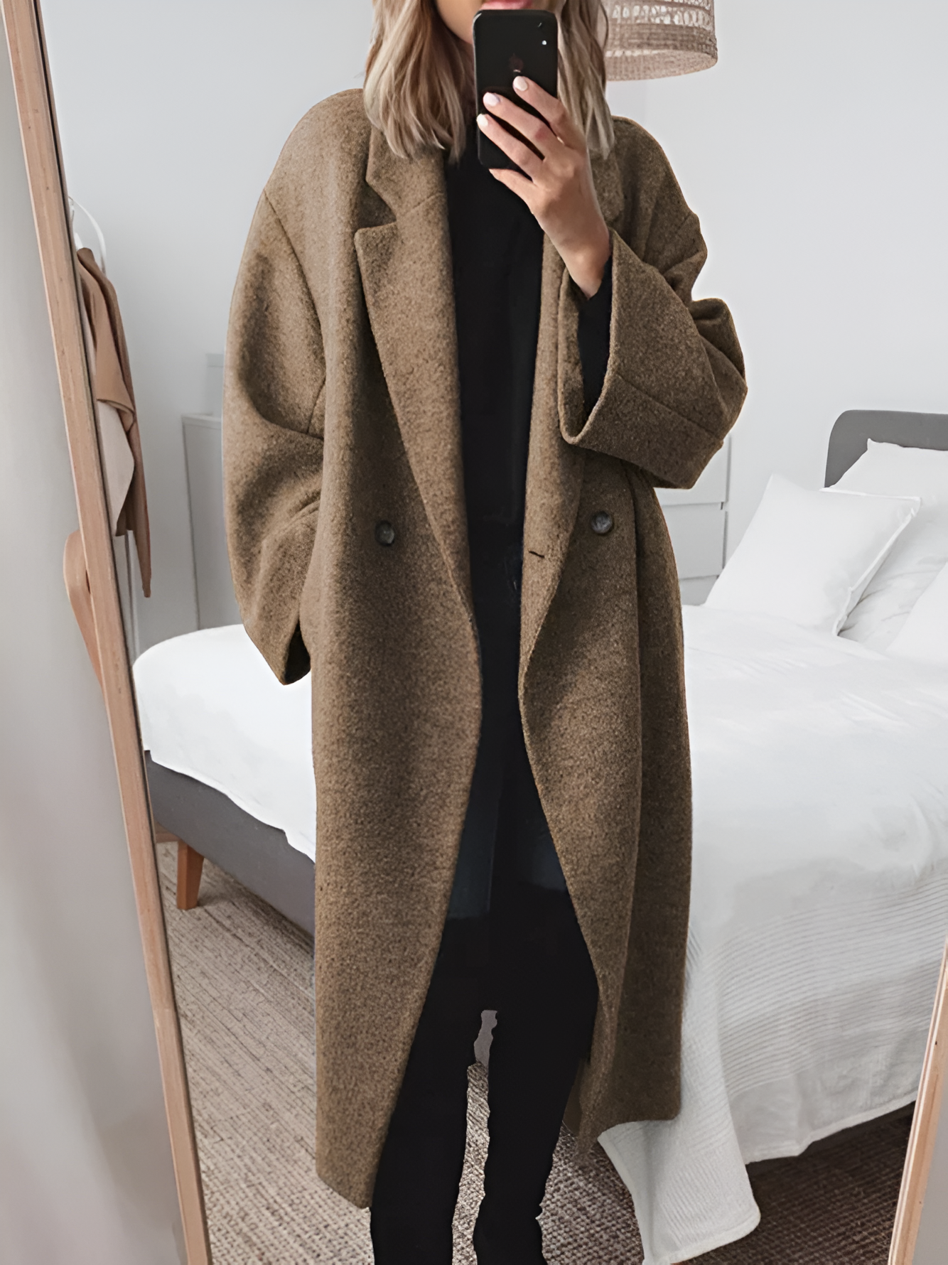 Femme en manteau oversize marron, tenant un smartphone devant un miroir, dans une chambre moderne. Mode automne-hiver, style décontracté.