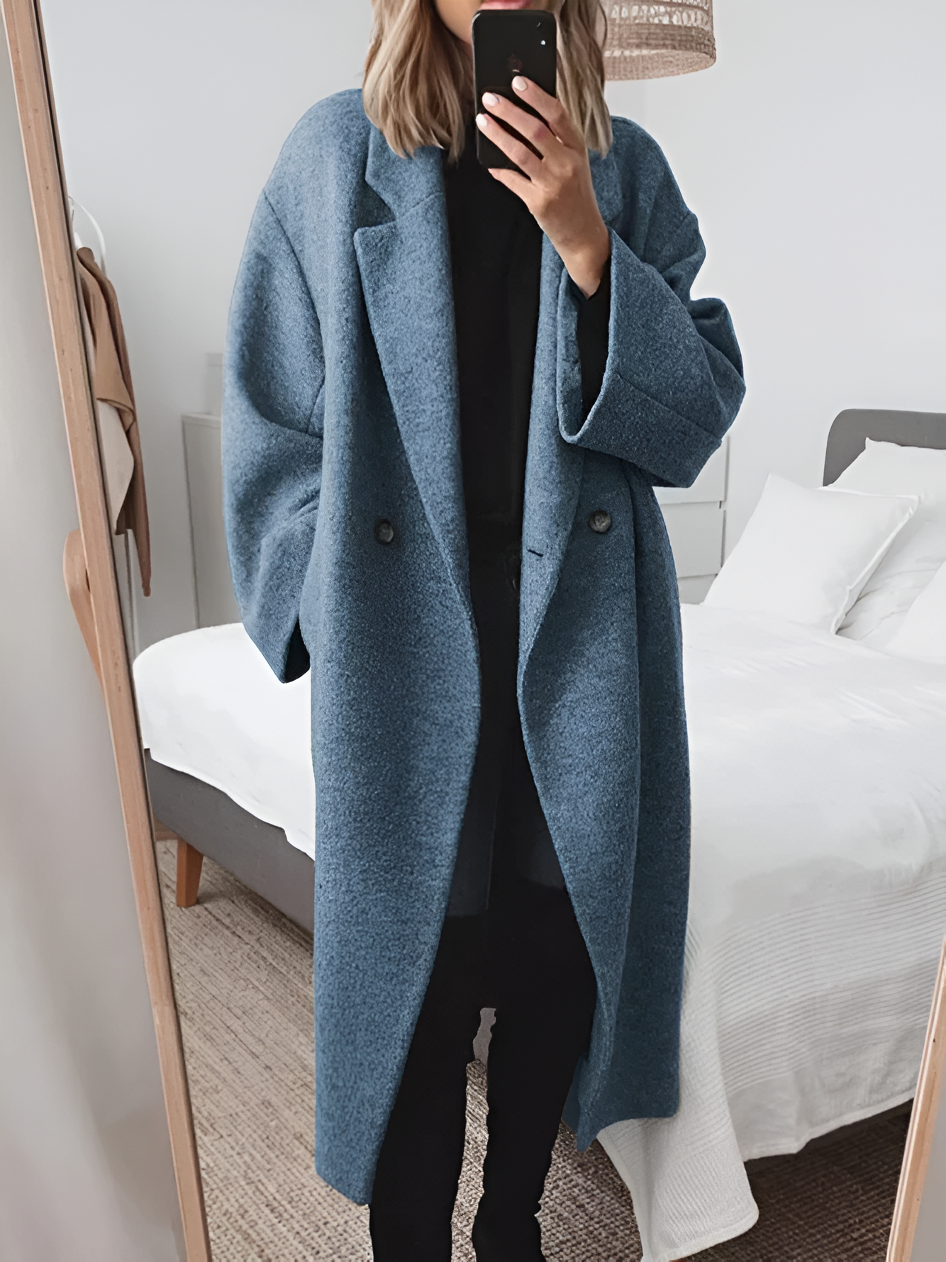 Femme en manteau bleu oversize, se prenant en photo dans un miroir, chambre moderne en arrière-plan, style mode automne-hiver tendance.