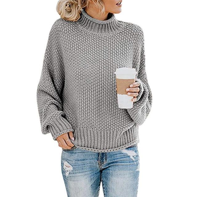 Femme portant un pull gris en maille avec un col roulé, tenant un gobelet de café, vêtue d'un jean déchiré. Mode décontractée automne-hiver.