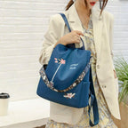 Femme portant un sac à dos bleu avec motifs floraux, tenue élégante beige, mur en briques blanches en arrière-plan. Mode féminine, accessoires tendance.