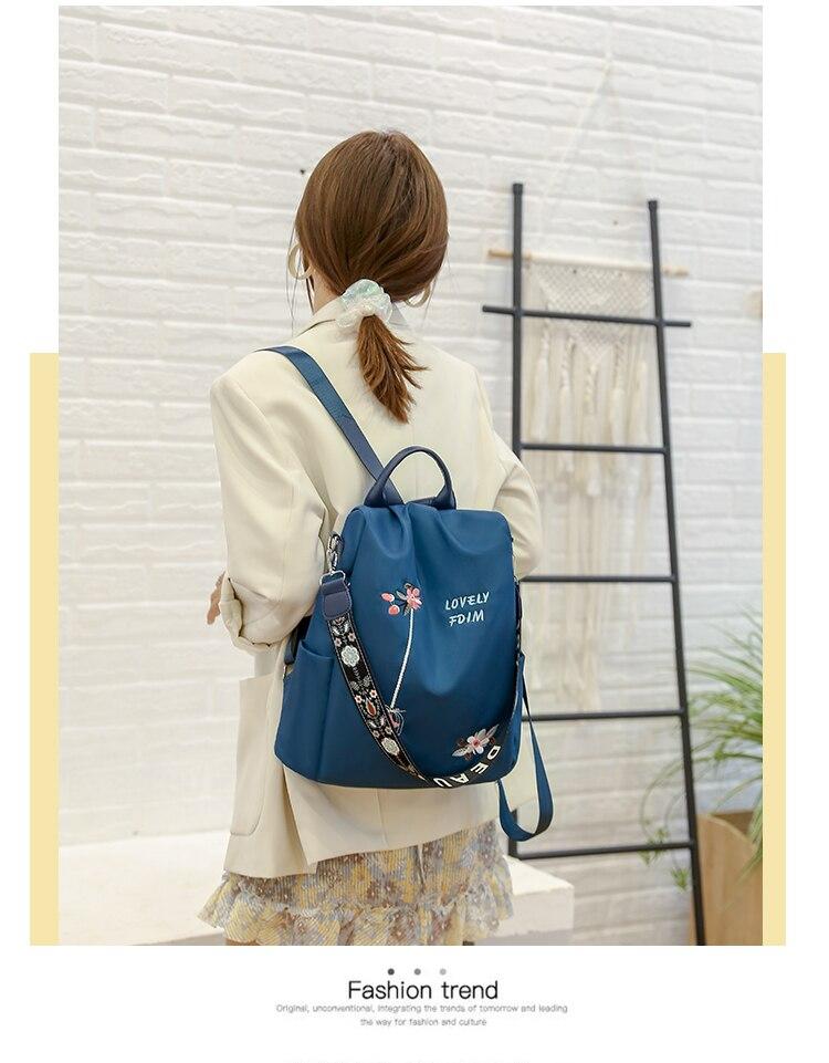 Femme de dos portant un sac à dos bleu avec motifs floraux, veste beige, robe à volants, devant un mur blanc et une échelle décorative.