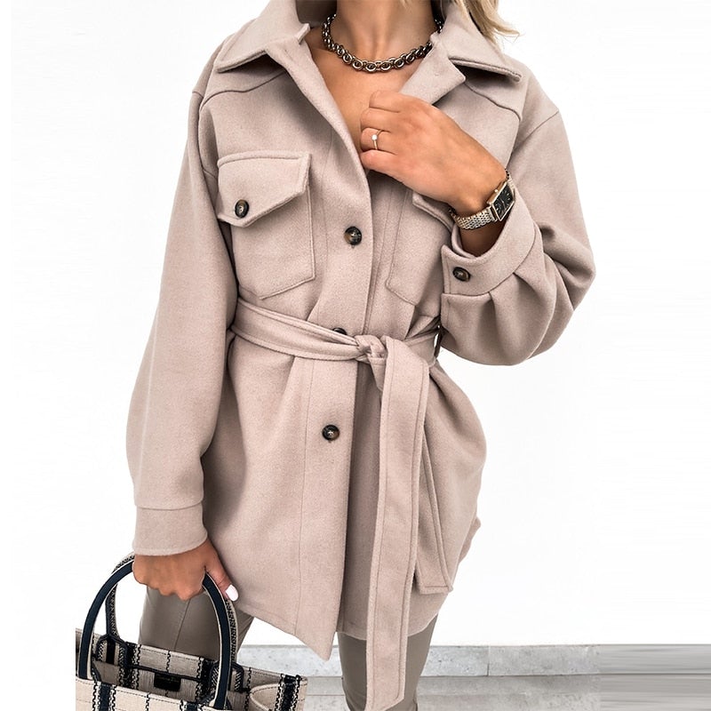 Femme portant un manteau beige ceinturé avec poches, accessoirisé d'un sac à main et d'une montre. Mode automne-hiver élégante et tendance.
