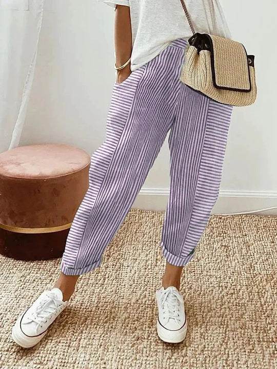 Pantalon rayé violet et blanc, style décontracté, porté avec un t-shirt blanc, baskets blanches et sac en osier, sur fond de tapis en jute.