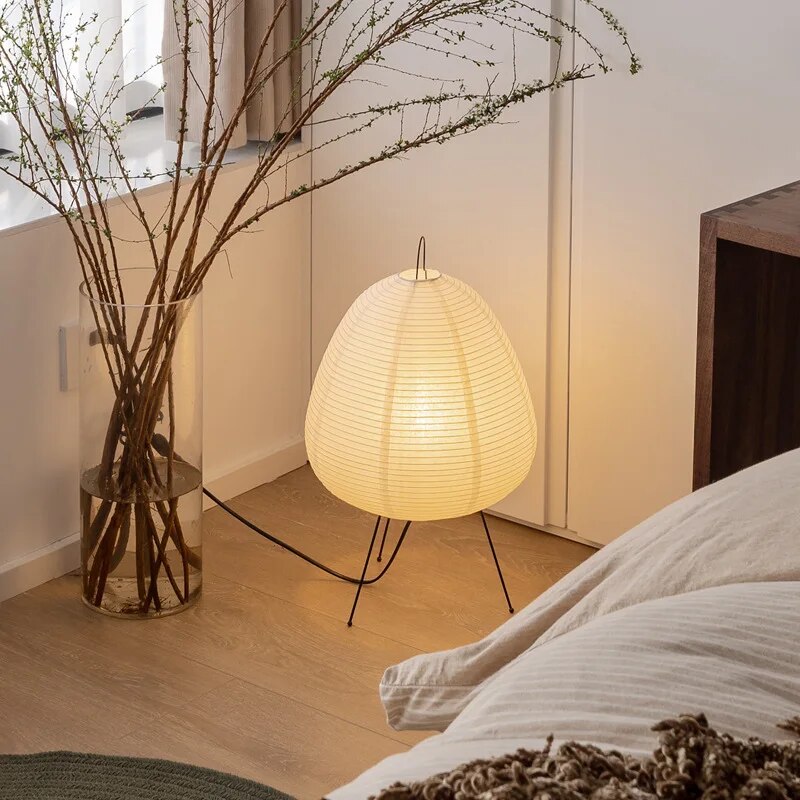 Lampe en papier japonaise allumée sur pied en métal, posée près d'un vase en verre avec des branches, dans une chambre moderne et minimaliste.