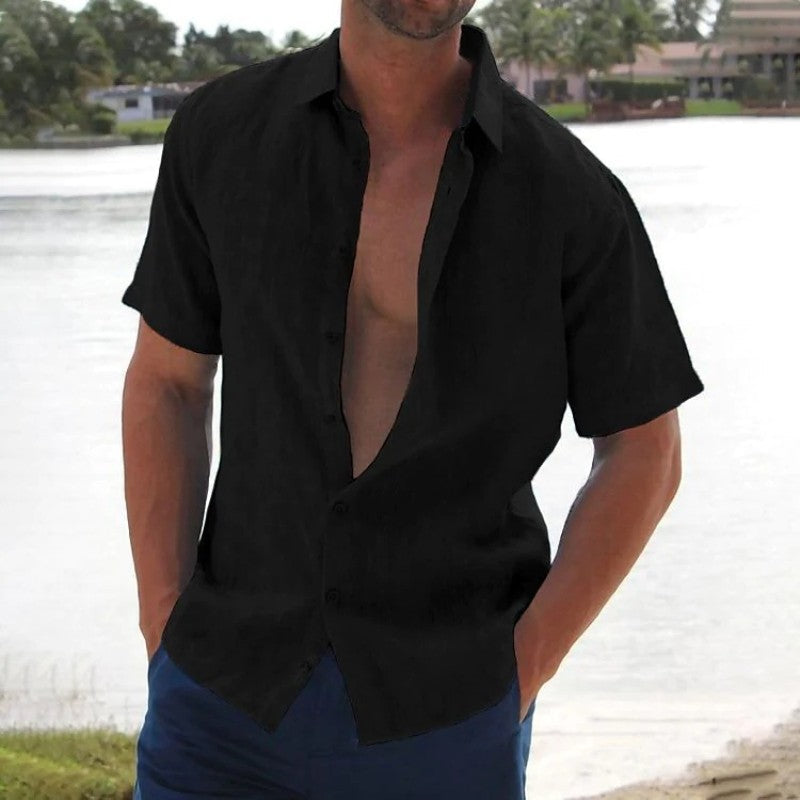 Homme en chemise noire ouverte, debout près d'un lac, paysage tropical en arrière-plan. Mode estivale décontractée, style masculin tendance.