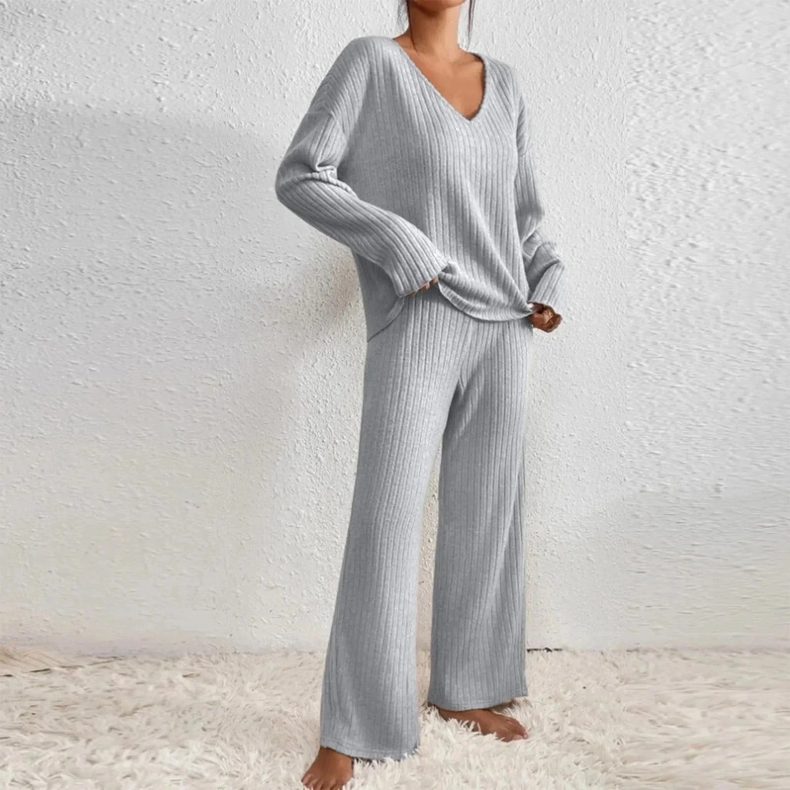 Femme portant un ensemble loungewear gris côtelé, composé d'un pull à col en V et d'un pantalon ample, sur un tapis blanc. Mode détente élégante.