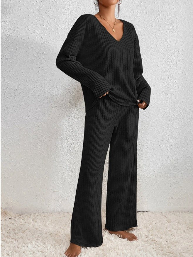 Femme portant un ensemble lounge noir en tricot côtelé, composé d'un pull à col en V et d'un pantalon ample, sur un tapis blanc.