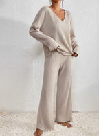 Femme portant un ensemble lounge en tricot beige, pull à col en V et pantalon ample, sur un tapis blanc. Mode confortable et élégante.