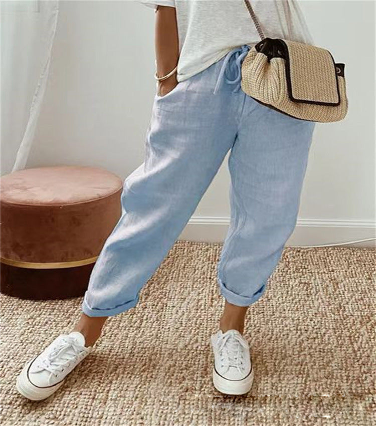 Femme portant un pantalon en lin bleu clair, t-shirt blanc, baskets blanches et sac en osier. Mode décontractée, tendance estivale, style chic.