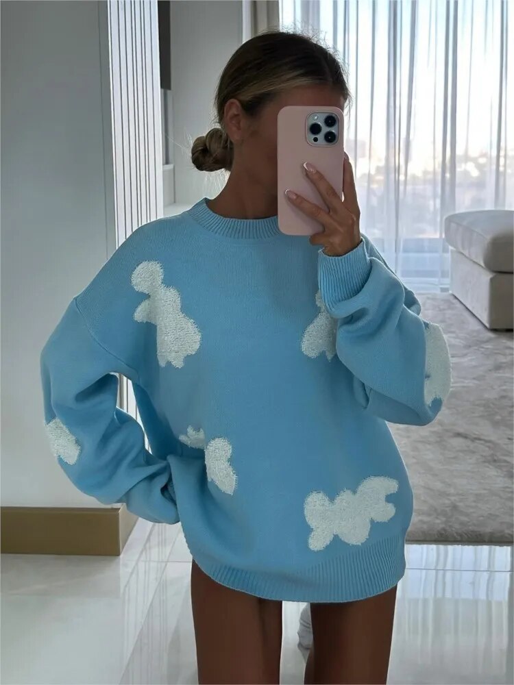 Femme portant un pull bleu avec motifs nuages blancs, tenant un smartphone rose, dans un salon moderne lumineux. Mode tendance, style décontracté.