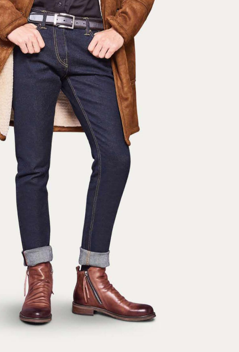 Homme portant un manteau marron, jean slim retroussé et bottines en cuir marron avec fermeture éclair, mode automne-hiver élégante et moderne.