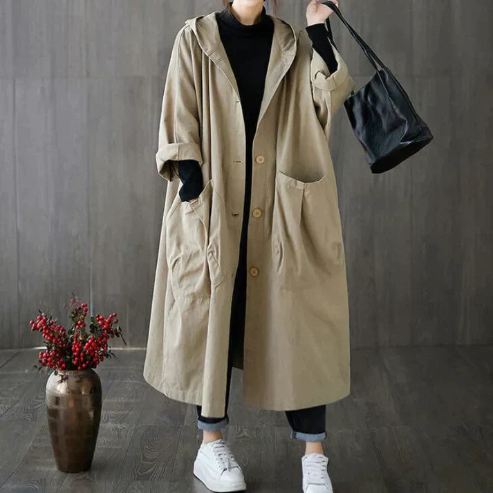 Manteau long beige à capuche, poches avant, porté avec un sac noir et des baskets blanches, sur fond de mur gris et vase décoratif.