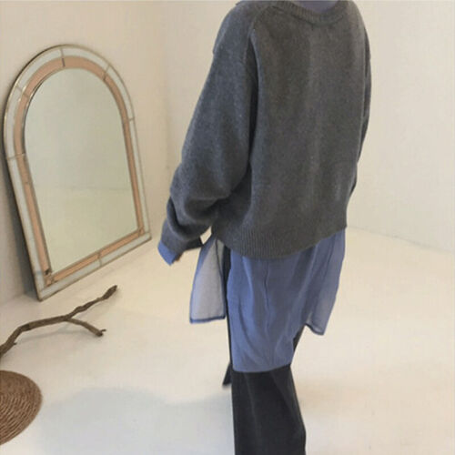 Personne portant un pull gris sur une chemise bleue, debout près d'un miroir ovale et d'une branche décorative. Mode décontractée, intérieur minimaliste.