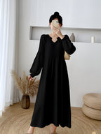 Femme en robe noire longue à manches longues, tenant un smartphone, dans un intérieur moderne avec plantes et décoration minimaliste.