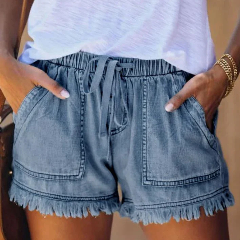 Short en jean bleu avec poches avant, taille élastique et ourlet à franges, porté avec un t-shirt blanc. Mode estivale décontractée pour femmes.