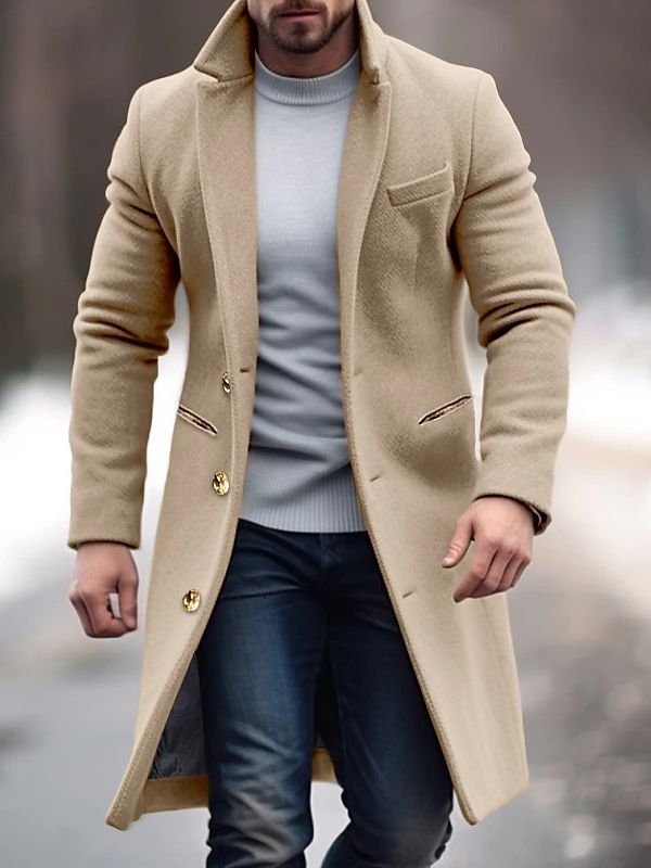Homme portant un manteau beige élégant, pull gris et jean, marchant en extérieur sur fond flou hivernal. Mode masculine, style hiver chic.