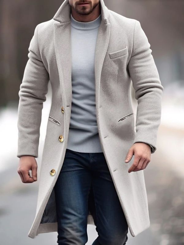 Homme portant un manteau long beige élégant, boutons dorés, sur un pull gris et un jean, marchant en extérieur. Mode masculine hiver chic.