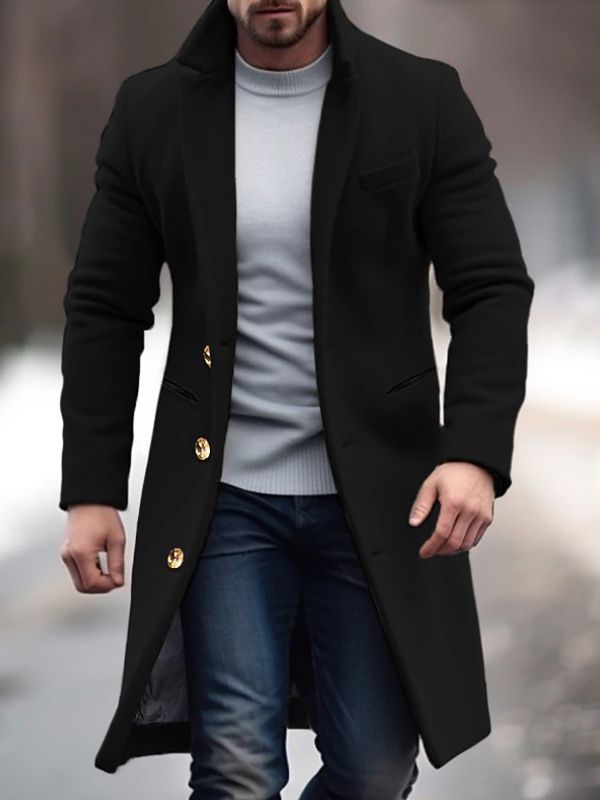 Homme portant un manteau noir élégant avec boutons dorés, pull gris et jean, marchant en extérieur sur fond flou hivernal. Mode masculine chic.