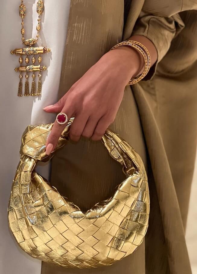 Sac à main doré tressé, tenu par une main ornée de bijoux élégants, incluant un anneau avec pierre rouge et bracelets dorés. Mode luxe et accessoires.