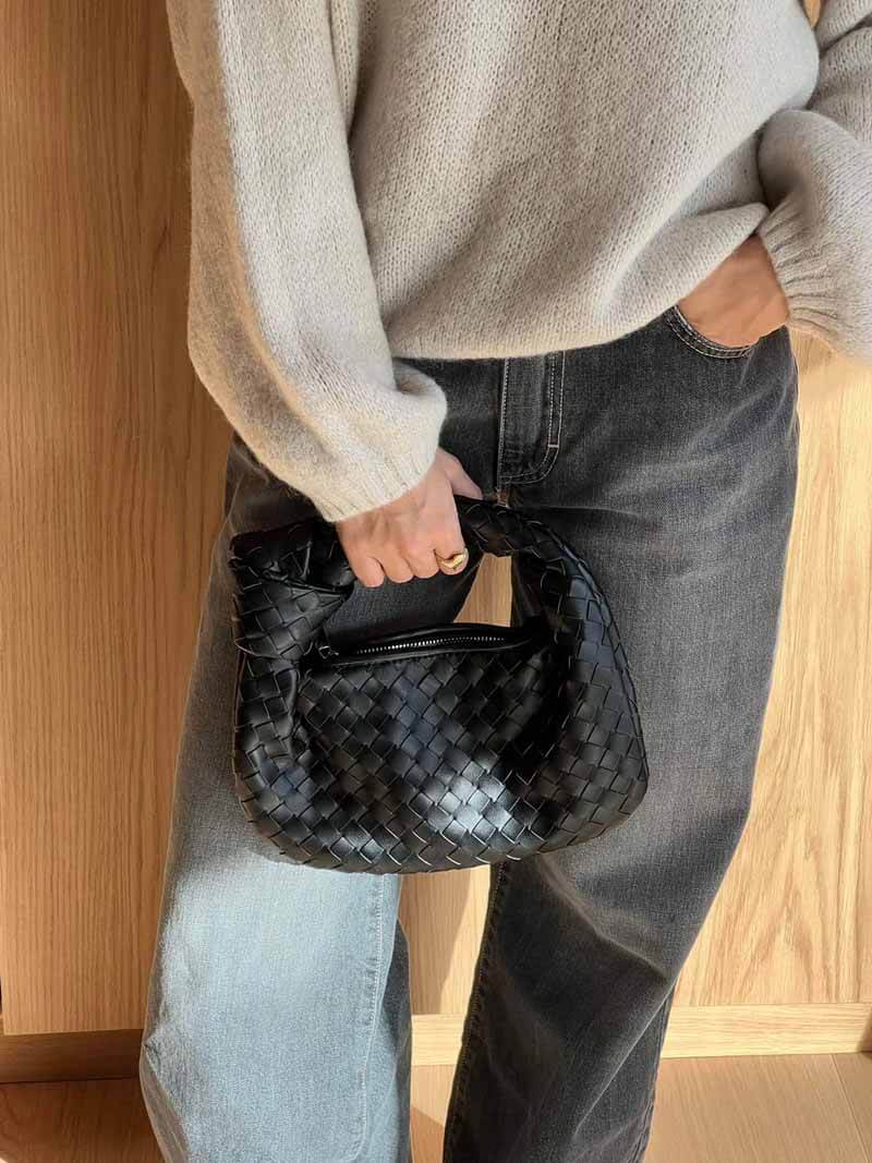 Sac à main noir tressé en cuir, porté par une personne en pull beige et jean gris. Mode élégante, accessoire tendance, style automne-hiver.