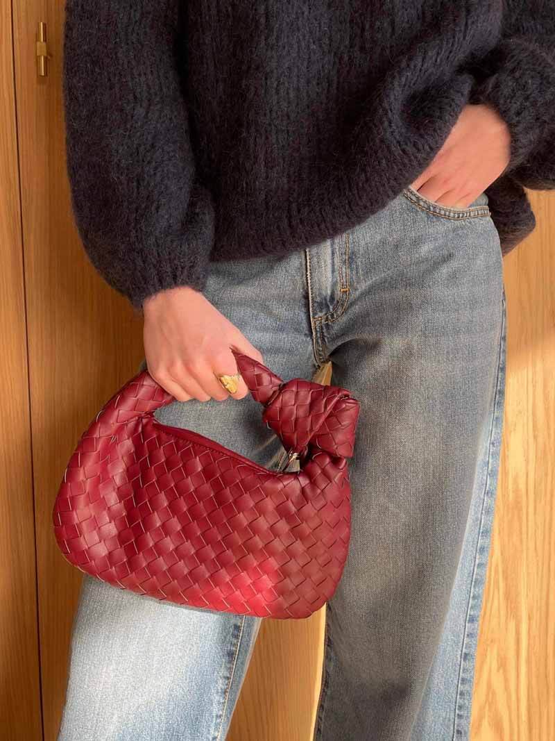 Sac à main rouge tressé en cuir, tenu par une personne en pull noir et jean. Mode élégante, accessoire tendance, style automne-hiver.