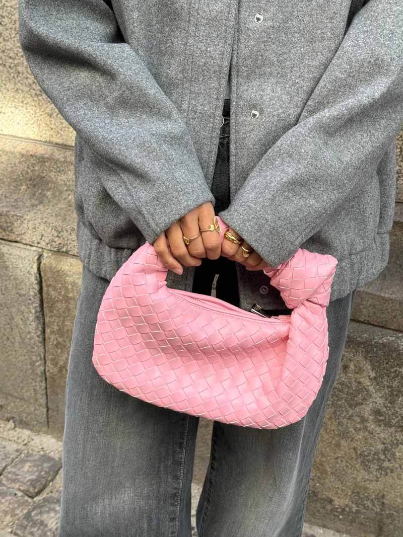 Personne tenant un sac à main rose tressé, vêtue d'un manteau gris et d'un jean. Mode urbaine, accessoires tendance, style décontracté.