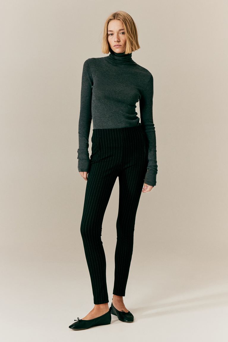 Femme portant un pull à col roulé gris et un pantalon noir à rayures, posant debout. Mode automne-hiver, tenue élégante et décontractée.
