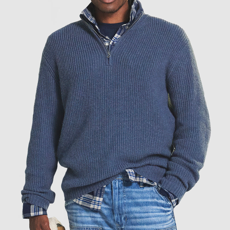 Homme portant un pull bleu à col zippé sur une chemise à carreaux, avec un jean décontracté. Mode masculine automne-hiver, style décontracté.