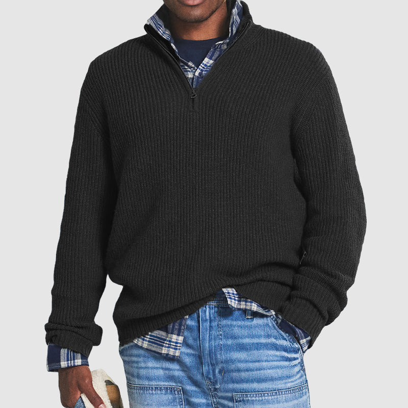 Homme portant un pull noir à col zippé sur une chemise à carreaux bleue, avec un jean décontracté. Mode masculine automne-hiver, style décontracté.
