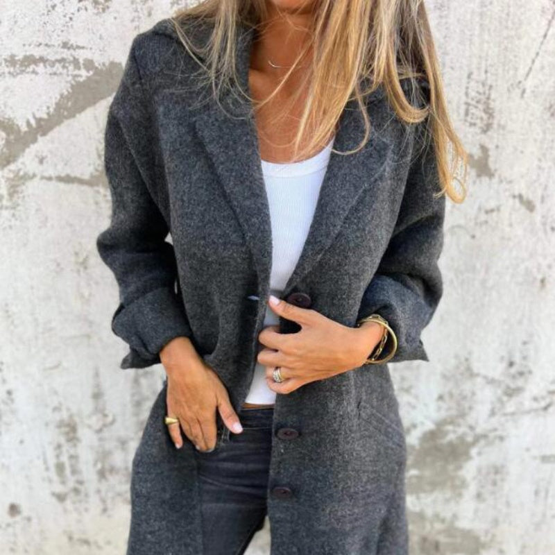 Femme portant un manteau gris élégant, chemise blanche et jeans noirs devant un mur texturé. Mode automne, style décontracté chic.
