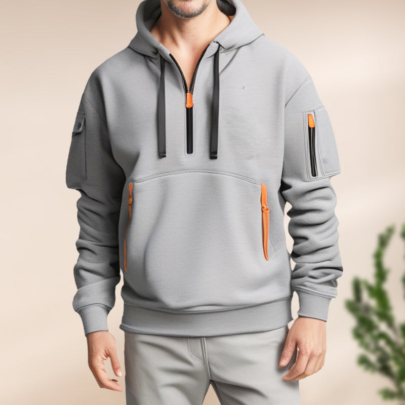 Homme portant un sweat à capuche gris moderne avec poches zippées orange, style décontracté, mode masculine tendance, fond neutre.