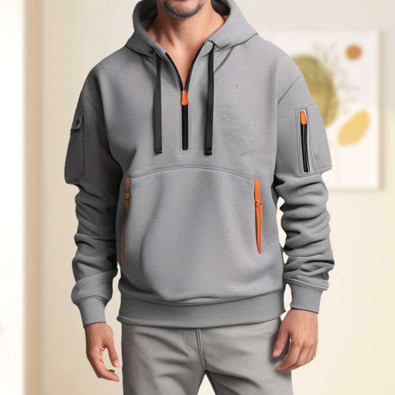 Homme portant un sweat à capuche gris moderne avec fermetures éclair orange, poches multiples, dans un intérieur lumineux. Mode décontractée tendance.