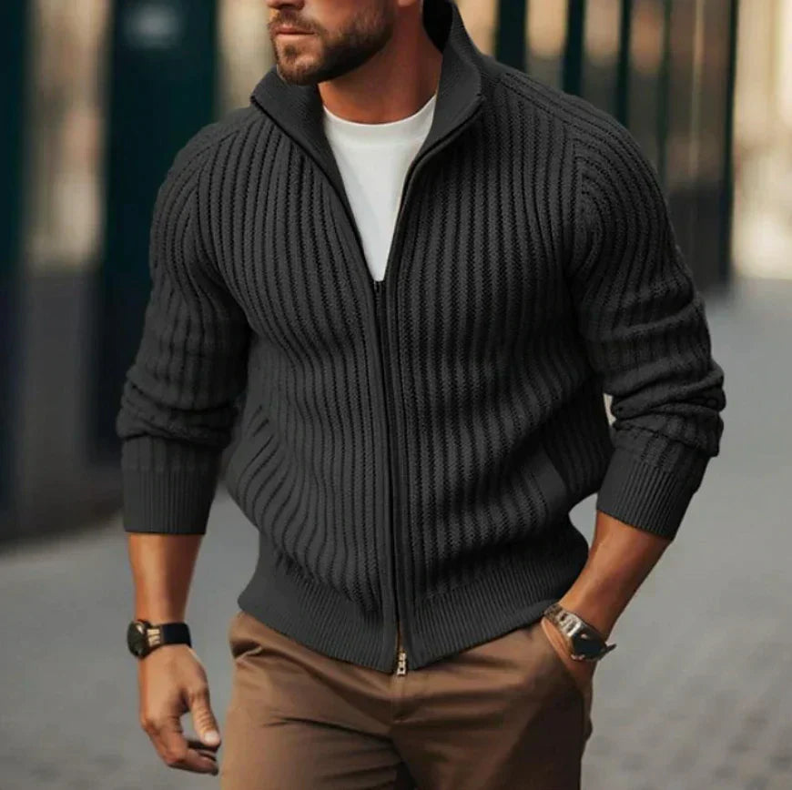 Homme portant un pull zippé en maille côtelée noire, t-shirt blanc, pantalon marron, montre noire, marchant en extérieur urbain. Mode masculine élégante.