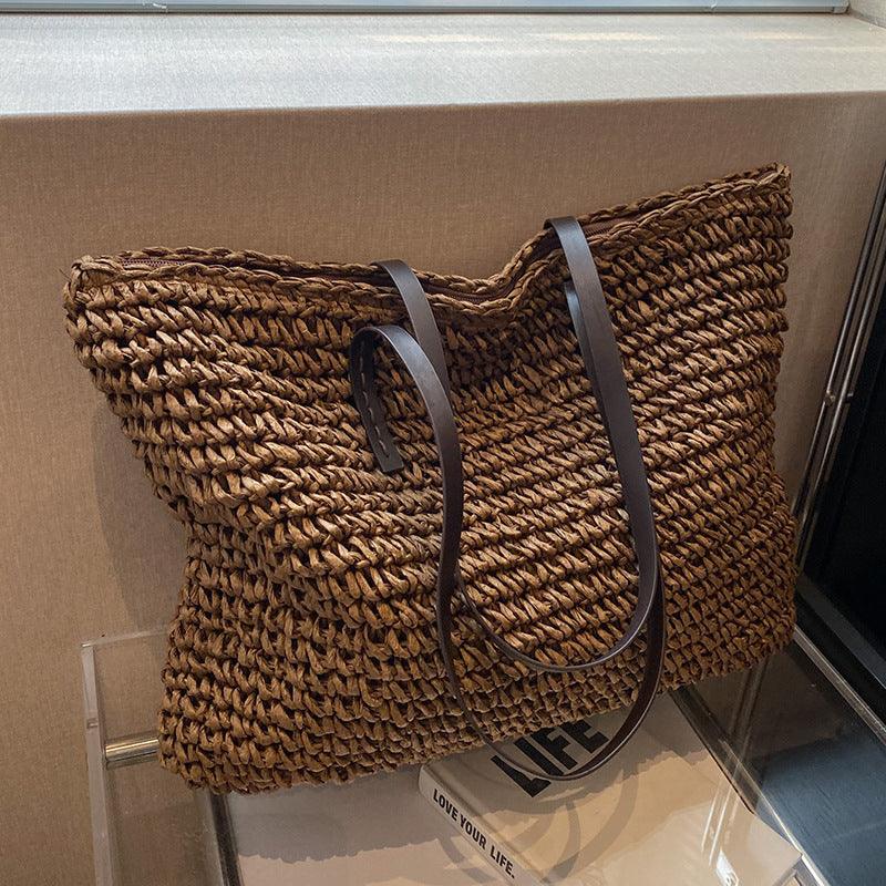 Sac cabas en paille tressée marron avec anses en cuir noir, posé sur une surface beige. Accessoire mode tendance, idéal pour l'été et le shopping.