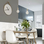 Salle à manger moderne avec table en bois, chaises blanches, horloge murale design, cuisine grise en arrière-plan, et suspension lumineuse.