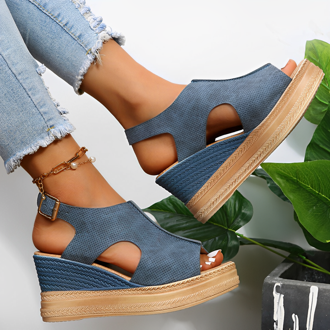 Sandales compensées bleues en toile, portées avec un jean déchiré, posées sur fond de feuilles vertes. Mode estivale, chaussures tendance femme.