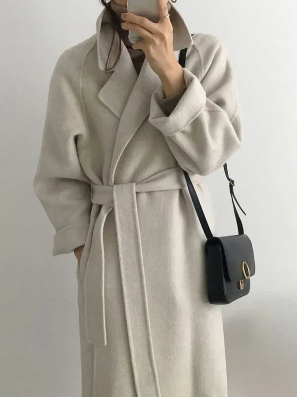 Personne portant un manteau beige élégant avec ceinture, tenant un smartphone, sac à bandoulière noir. Mode automne-hiver, style chic et moderne.