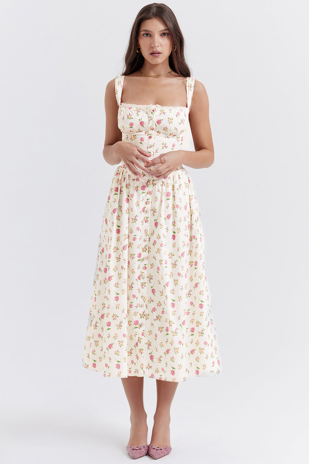Femme portant une robe longue à fleurs, fond blanc, style vintage, bretelles larges, élégante et estivale, chaussures roses, mode féminine chic.