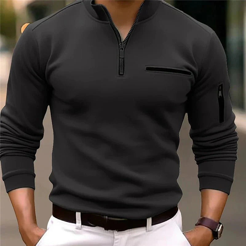 Homme portant un pull noir à col zippé, manches longues, avec poche poitrine, sur fond urbain flou. Mode masculine élégante et moderne.