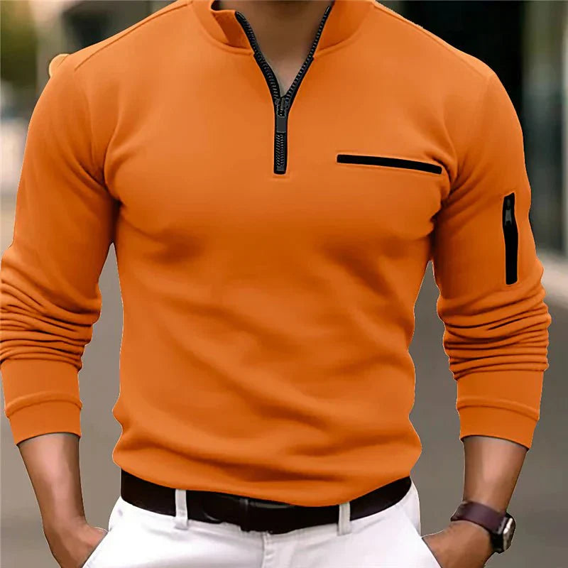 Homme portant un pull orange à col zippé, poche poitrine et manche, associé à un pantalon blanc. Mode masculine élégante et moderne.