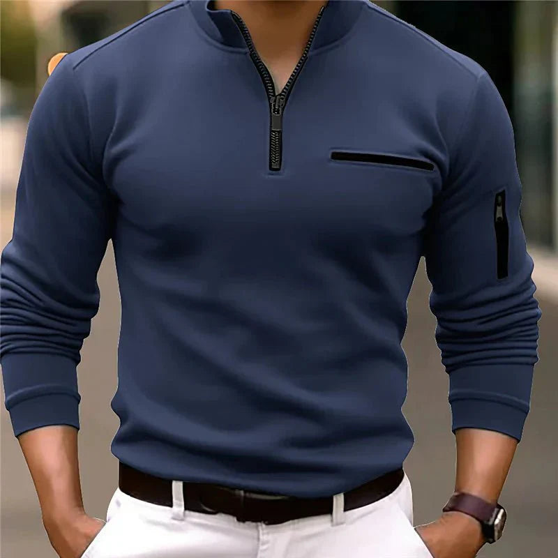 Homme portant un pull bleu à col zippé, manches longues, avec poche poitrine, associé à un pantalon blanc. Mode masculine élégante et moderne.