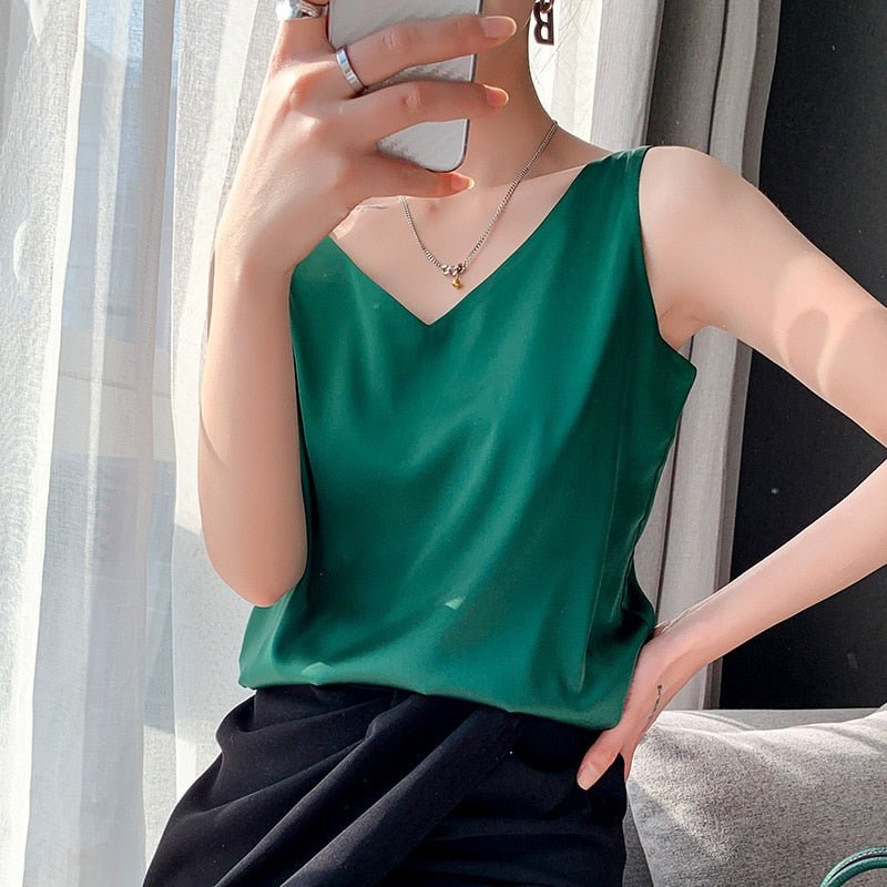 Femme portant un débardeur vert émeraude, tenant un smartphone, assise près d'une fenêtre. Mode élégante, bijoux minimalistes, lumière naturelle.