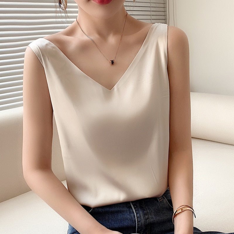 Femme portant un débardeur en satin blanc, col en V, assise sur un canapé beige, avec un collier délicat et un bracelet doré. Mode élégante et chic.