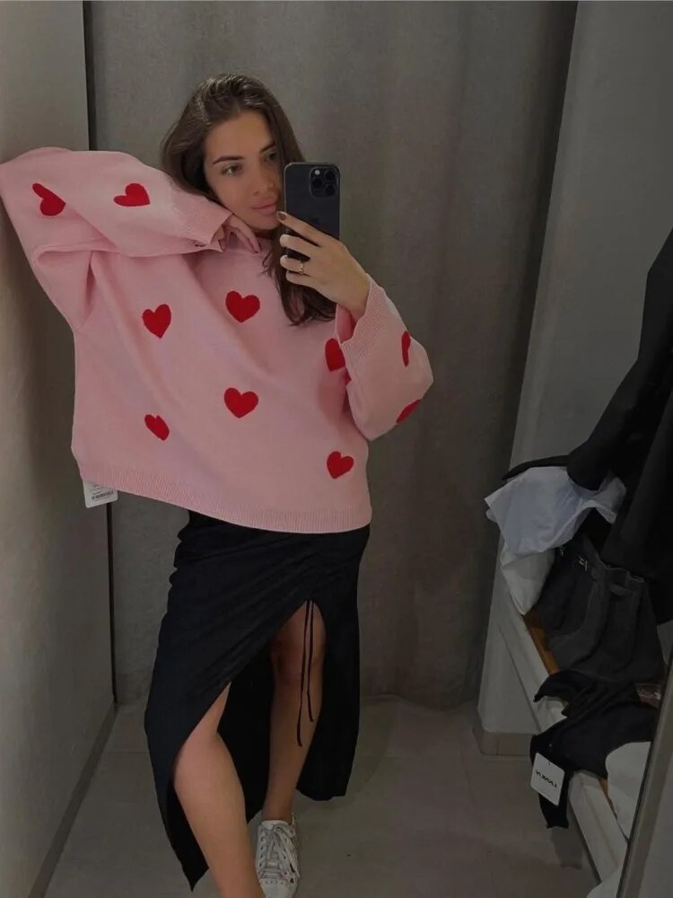 Jeune femme prenant un selfie dans un miroir, portant un pull rose à motifs de cœurs rouges et une jupe noire fendue, dans une cabine d'essayage.