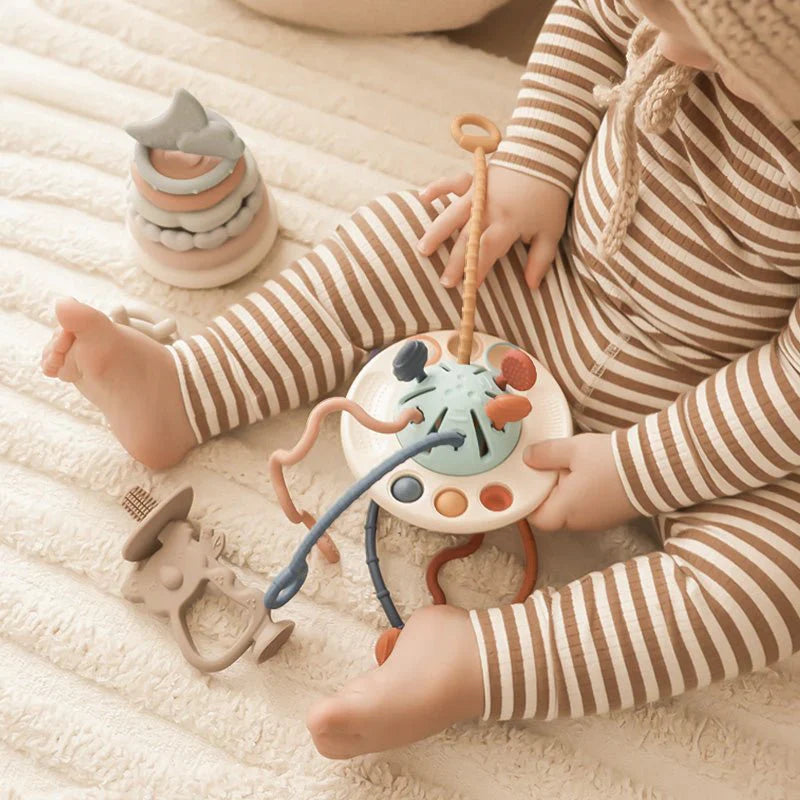 Bébé jouant avec jouet éducatif en silicone, anneaux empilables et hochet, sur couverture beige. Jouet sensoriel pour développement enfant.