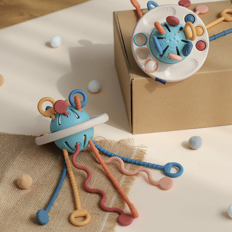 Jouet sensoriel pour bébé en silicone bleu avec anneaux et textures variées, posé sur une boîte en carton. Accessoire d'éveil Montessori.