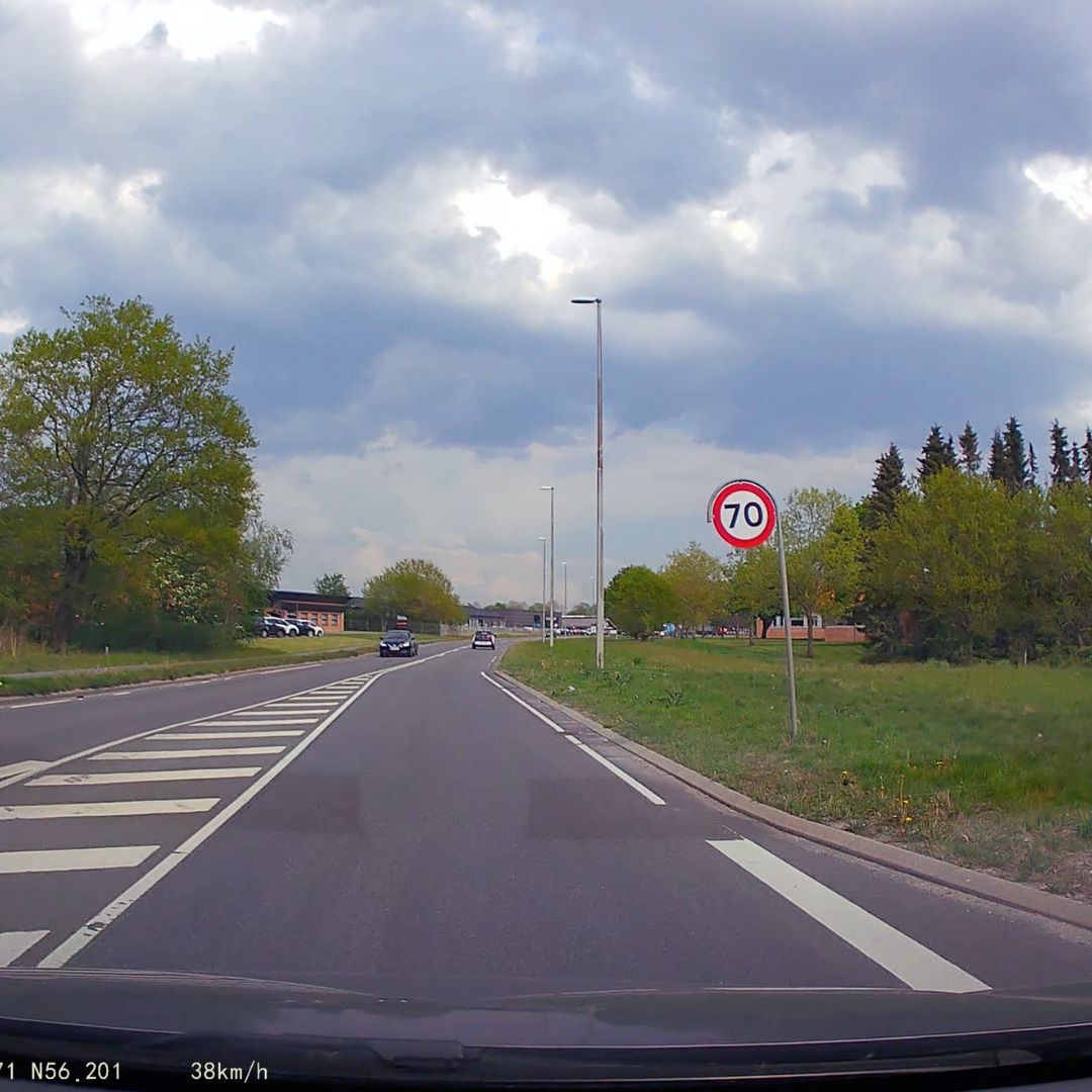 Route de campagne avec panneau de limitation de vitesse à 70 km/h, ciel nuageux, arbres verts, et voitures en circulation. Paysage printanier.