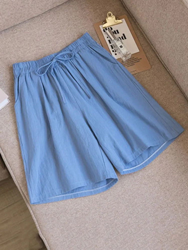 Short bleu en lin pour homme posé sur un canapé beige, avec une ceinture à cordon. Mode estivale, vêtement décontracté, style confortable.