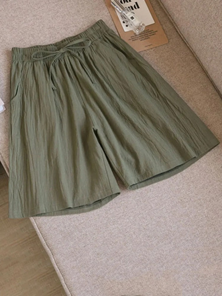 Short en lin vert olive sur canapé beige, style décontracté, avec cordon de serrage et poches latérales. Mode estivale, confort et élégance.